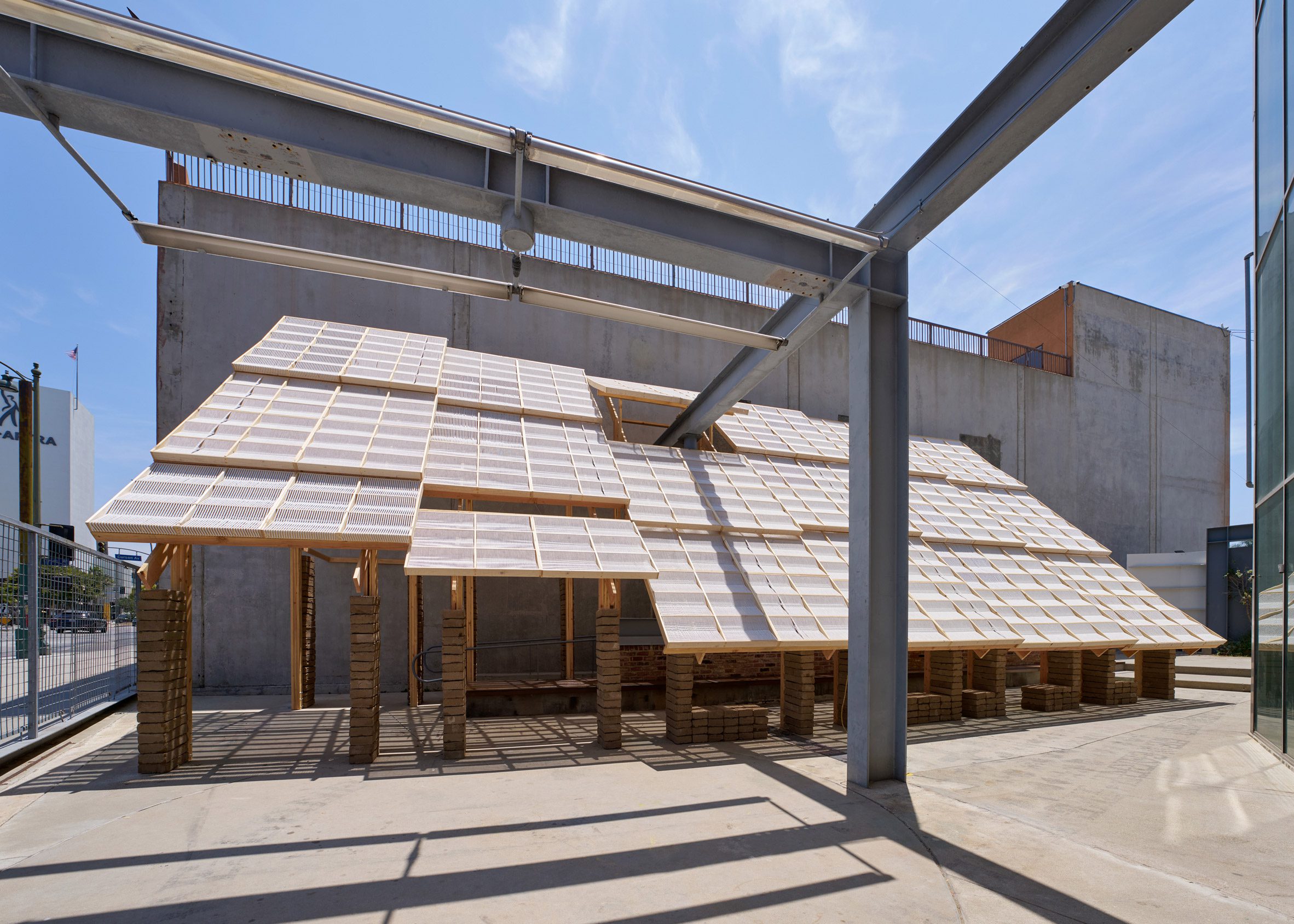 A view through the woven canopy and earthen columns of the Earthen Comforts pavilion, showing dappled shade